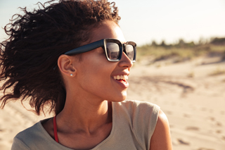Happy young woman with curly hair wearing sunglasses, smiling on a sunny beach day.