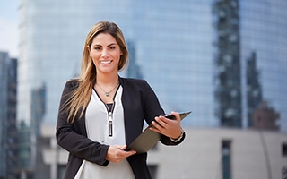 Professional woman in business attire holding a clipboard with a pen in front of a glass building.