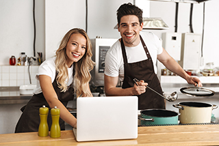 Man and woman in aprons cooking together and using laptop in modern kitchen.
