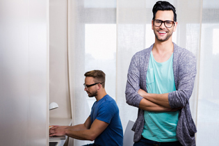 Two young men in an office, one sitting and working on a laptop, the other standing with arms crossed and smiling at the camera.
