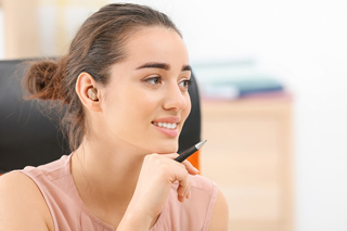 Young woman in office environment holding a pen, looking thoughtful with a slight smile.