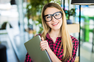 Young woman in glasses and plaid shirt holding a laptop in an office environment.