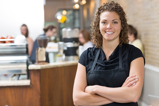 Woman with curly hair wearing a black apron standing in a café with arms crossed.