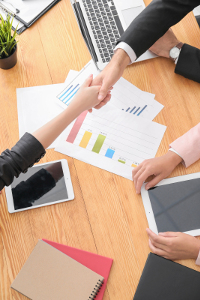 Two professionals shaking hands over a desk with business charts, laptop, and digital tablet during a meeting.