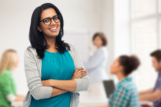 Confident woman in glasses standing with arms crossed in a classroom, smiling, with students and teacher blurred in the background.