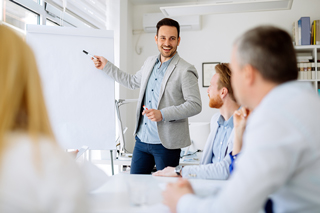 Businessman giving a presentation with a marker in a bright office setting, audience of colleagues listening attentively.