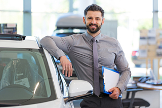 Confident car salesman standing with arm on a white car in showroom, holding a blue folder and smiling at the camera.
