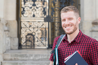 Male student with short hair, wearing a red plaid shirt and carrying a backpack and a book, standing in front of an ornate historical building door.