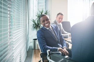 Two businessmen smiling in an office setting, one in the foreground in a wheelchair and another standing in the background by the window with blinds.