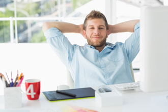 Relaxed young man with hands behind head smiling at desk with computer and office supplies in office setting.