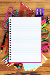 Blank spiral notebook surrounded by colorful school supplies including pens, pencils, scissors, rulers, and a sharpener on a wooden background