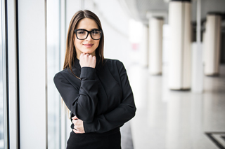 Professional woman in glasses wearing a black blazer standing in an office corridor