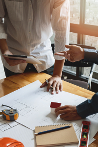 Two architects discussing over blueprints with digital tablet and construction tools on desk in a well-lit office setting.