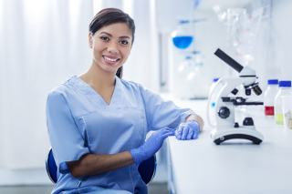 Female lab technician in blue scrubs sitting in a laboratory with a microscope and lab equipment in the background.