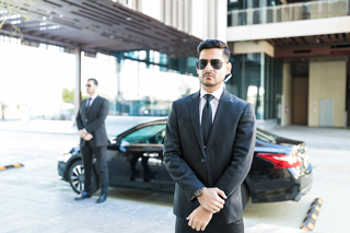 Professional male bodyguard wearing sunglasses and a suit standing in front of a luxury car with another bodyguard in the background at a hotel entrance.
