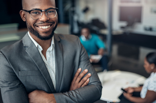 Confident African American businessman smiling in office, wearing glasses and a gray suit, with colleagues working in background.