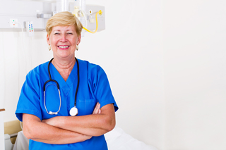 Smiling elderly nurse with blonde hair wearing blue scrubs and a stethoscope around her neck, standing with arms crossed in a hospital room.