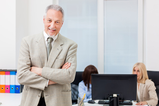 Senior businessman with arms crossed smiling in office environment with female colleagues working in background.