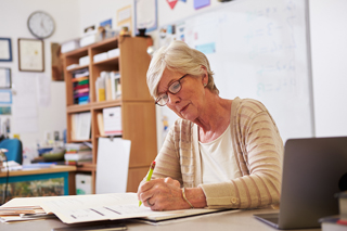 Elderly female teacher grading papers at her desk in a classroom, laptop beside her, with a whiteboard in the background.