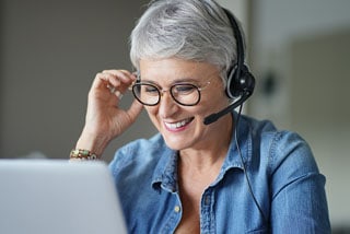 Smiling senior woman with gray hair wearing glasses and a headset using a laptop in an office setting.