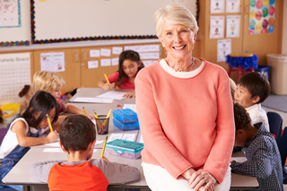 Smiling senior female teacher sitting in front of a classroom with diverse students engaged in activities, educational posters on walls.