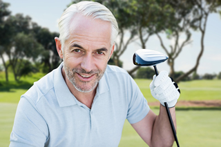 Senior man with gray hair smiling, holding a golf club on a golf course with trees in the background.