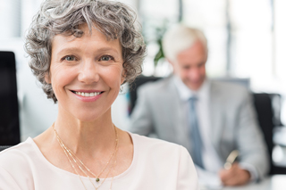 Smiling senior woman with curly gray hair wearing a pink blouse in an office setting, with an older man working in the background