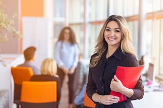 Professional woman in business attire smiling with a red folder in an office environment with colleagues in background.
