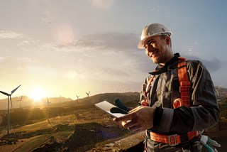 Engineer in hard hat and safety harness holding documents at wind farm during sunset