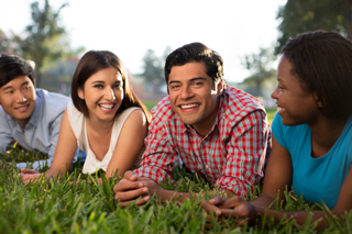 Four young adults lying on grass and smiling, enjoying a sunny day outdoors, with focus on multiethnic friendship and leisure.