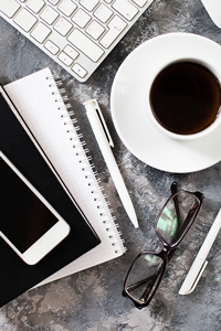 Top view of a modern workspace with coffee cup, smartphone, notebook, pen, glasses, and keyboard on a textured gray surface.