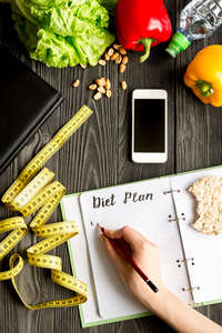 Person writing a diet plan in a notebook surrounded by healthy foods including vegetables, nuts, and a measuring tape, with a smartphone on a wooden table.