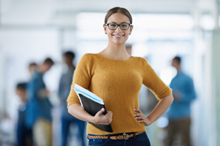 Confident young woman wearing glasses and a mustard sweater, holding books in a busy office environment.