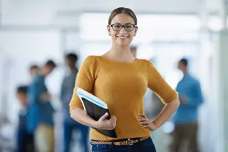 Confident businesswoman with glasses holding a notebook in an office environment, smiling at the camera with colleagues in the background