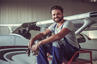 A cheerful airplane mechanic sitting on a ladder in front of a small aircraft inside a hangar, holding a cleaning cloth and wearing work overalls.