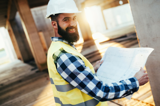 Construction worker with beard wearing hard hat and reflective vest holding blueprints at a construction site.