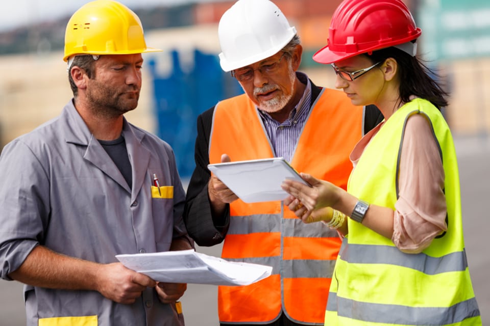  Three people wearing hard hats and safety vests are looking at a tablet. The image represents the search query 'Jobs that provide housing'.