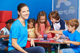 Young female teacher and diverse group of children engaged in arts and crafts activities at a classroom table, smiling and focused on their projects.