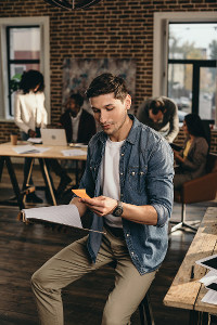Young man in casual attire reviewing documents in a bustling office with brick walls and coworkers in background.