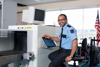 Security officer operating an X-ray machine at an airport checkpoint, American flag in background.