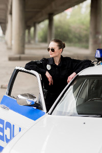 Female police officer in sunglasses leaning on a patrol car under a bridge