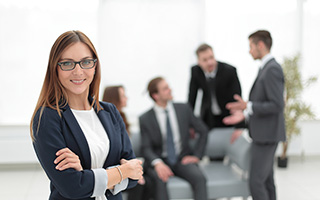Professional woman with glasses smiling in foreground, three male colleagues conversing in background in office setting.