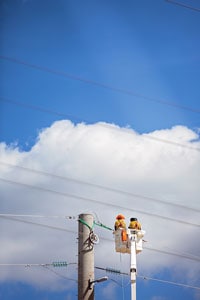 Two utility workers in protective gear repairing power lines from a cherry picker against a blue sky with clouds