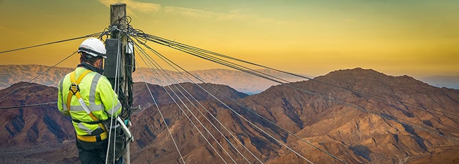 A lineworker positioned near the top of a utility pole overlooking a mountain range