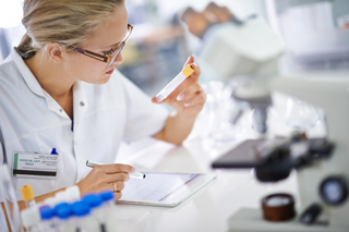 Female scientist examining a test tube in a laboratory while taking notes on a digital tablet.