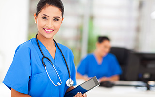 Nurse in blue scrubs with stethoscope holding a tablet in hospital setting with another healthcare professional in the background