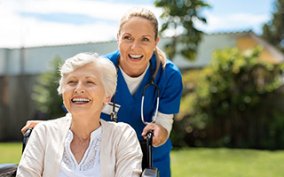 Elderly woman in wheelchair with caretaker smiling outdoors in sunny garden