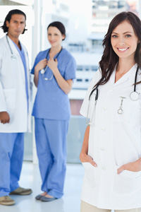 Three healthcare professionals in a hospital setting, featuring a Woman in a white coat in the foreground, and a man and woman in blue scrubs in the background.