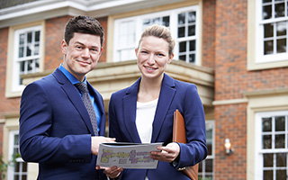 Two real estate agents holding house plans in front of a brick residential building.