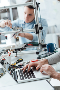 Two people working in a technology lab, one adjusting elements on a 3D printer while the other uses a laptop, displaying teamwork and innovation in a modern workspace.
